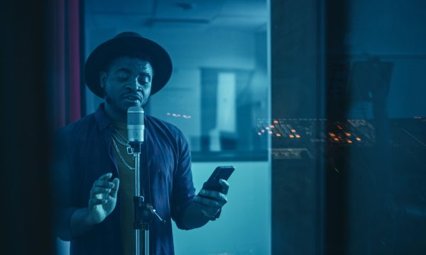 Portrait of Successful Young Black Artist, Singer, Performer Singing His Hit Song for the New Album. Wearing Stylish Hat, Holding Smartphone and Standing in Music Record Studio Soundproof Room.