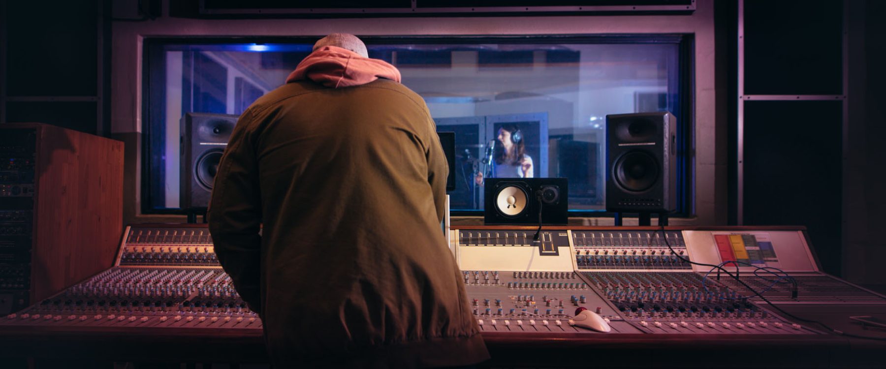 Sound engineer working at audio control panel with singer singing in recording room in background. Musicians producing music in professional recording studio.